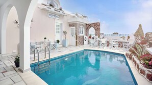 A pool area with a clear blue pool, white outdoor furniture, and a stone wall with a door. - Merovigliosso Budget Studio ground floor (Thira - Santorini)