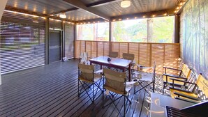 Un espacio de estar techado al aire libre con un suelo de madera, una mesa con sillas y luces colgantes.