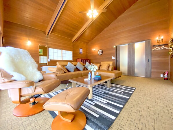 A spacious living room with a high ceiling, wooden walls, a sofa set, a coffee table, and a patterned rug.