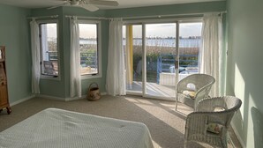 Ein Schlafzimmer mit Bett, Sessel, Deckenventilator und Blick auf ein Gewässer durch große Schiebetüren.