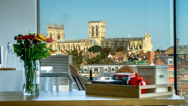 A view of a historic cathedral through a window, with a vase of flowers and a biscuit box on the windowsill. - Cityscape @ Ryedale House -  a family break that sleeps 4 guests  in 2 bedrooms (York)