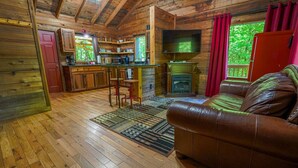 Un interior de cabaña acogedor con suelo de madera, un sofá de cuero, una zona de cocina con televisor y una chimenea.