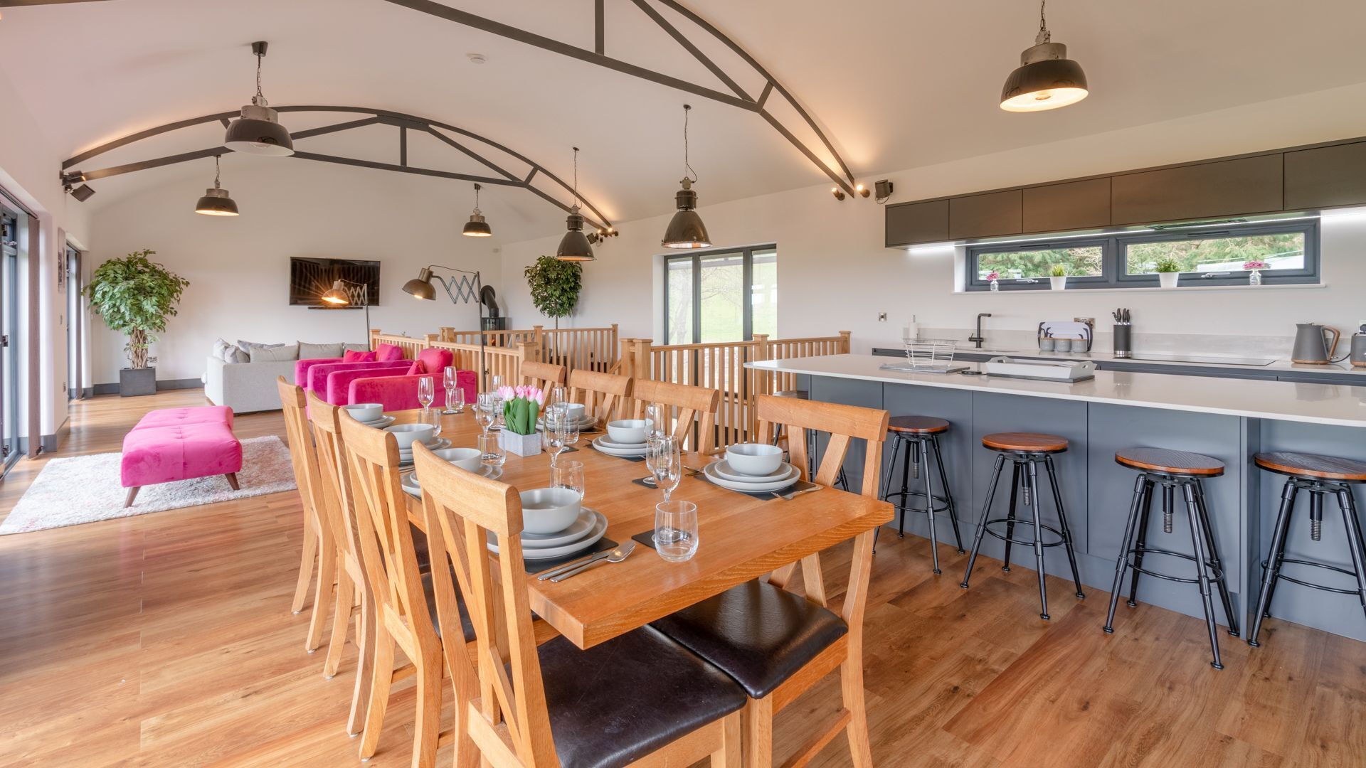 Een ruime eetruimte met houten tafels en stoelen, een keuken met een groot raam, en een gezellige zithoek met een roze poef.