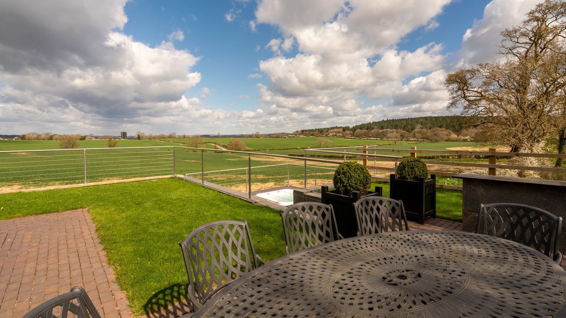 A patio with a metal table and chairs, a view of a green field, and a cloudy sky.