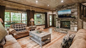 A cozy living room with a stone fireplace, wooden ceiling, and large windows offering a view of the outdoors. - Luxury, Cozy & Fun ★ Pool, Hot Tub, Theater, Games (Sevierville)