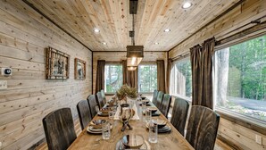 A wooden-paneled dining room with a long table set for six, large windows, and framed artwork on the walls. - Luxury, Cozy & Fun ★ Pool, Hot Tub, Theater, Games (Sevierville)