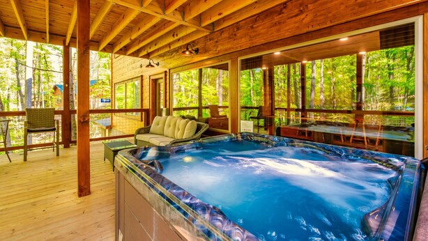 A wooden deck with a hot tub, wicker furniture, and large windows overlooking a forest. - Luxury, Cozy & Fun ★ Pool, Hot Tub, Theater, Games (Sevierville)
