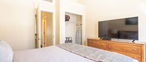 A bedroom with a bed, a television on a wooden dresser, a wardrobe, and a door leading to another room.