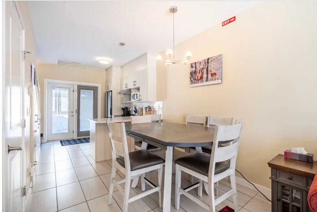 A dining area with a table and chairs, a kitchen in the background, and a door leading outside.