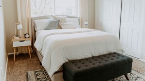 Une chambre à coucher comprenant un lit, des tables de chevet, une commode, une fenêtre avec des rideaux, une porte et un tapis à motifs.