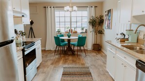 Une cuisine moderne avec des meubles blancs, une table à manger entourée de chaises turquoise, un plancher en bois et une grande fenêtre agrémentée de rideaux.