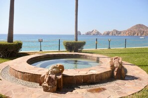 A circular stone pool with rock sculptures, surrounded by a rope barrier, with a view of the ocean and mountains in the background.
