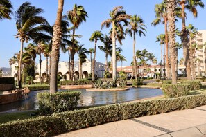 A landscaped area with palm trees, a water feature, and a building with arches in the background.