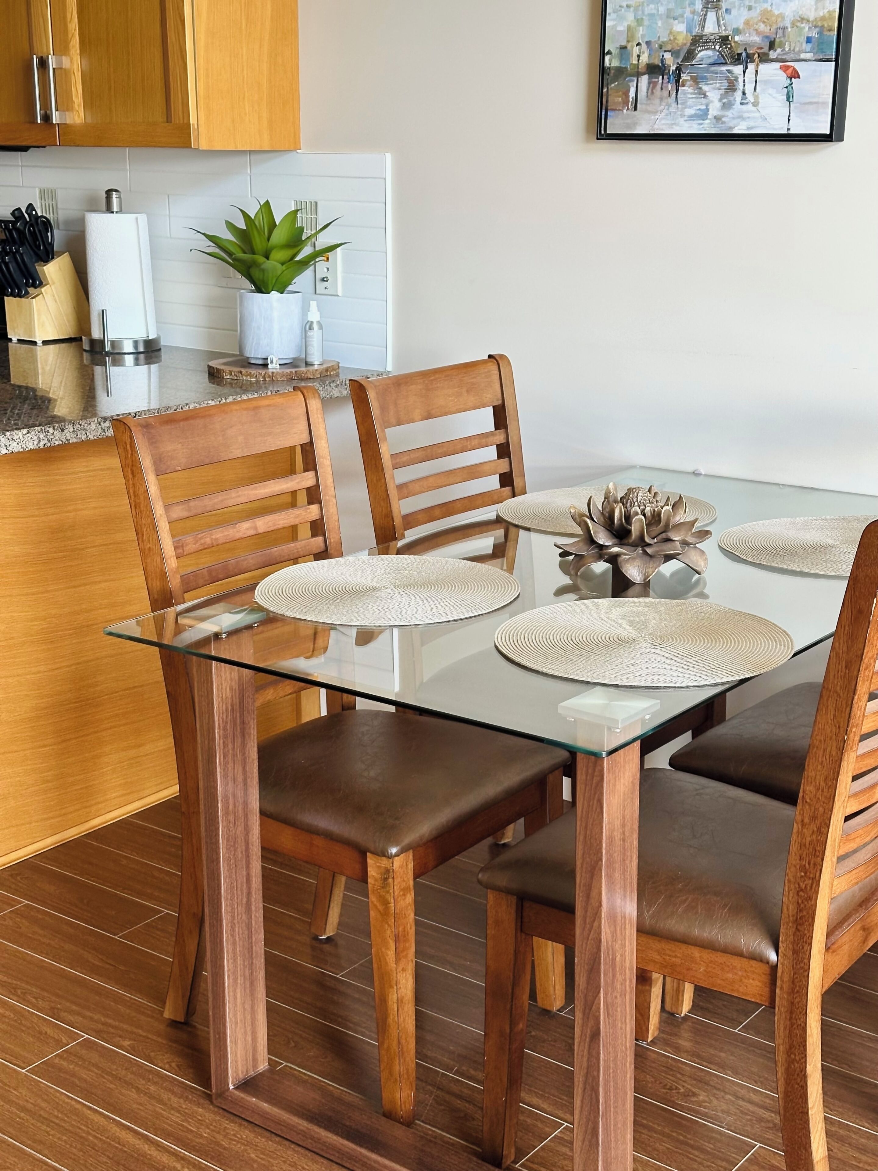 A modern kitchen with wooden cabinets, a glass dining table with four chairs, and a potted plant.