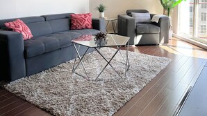 Un salon moderne avec un canapé foncé, une table basse en verre et une grande fenêtre donnant sur des immeubles.