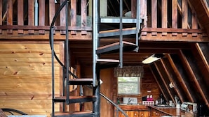 A rustic cabin interior with a spiral staircase leading to a loft bedroom, wooden floors, and a kitchen area.