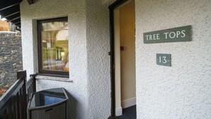 A building exterior with a textured wall, a window, a door labeled "TREE TOPS 13", and a small outdoor seating area. - Tree Tops at The Falls -  3-bedroom duplex, stroll into Ambleside (Ambleside)