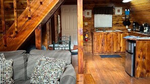 A cozy cabin interior with a wooden staircase, a gray sofa with patterned cushions, a kitchen area with a refrigerator, and a dining table.