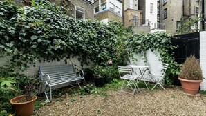 A courtyard with a gravel ground, a white bench, a white table, and potted plants. - 2 Bed 2 Bath Garden Flat in Victoria Pimlico (London)