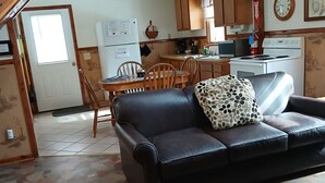 Una sala de estar acogedora con un sofá de cuero negro, una mesa de comedor con sillas y una zona de cocina con armarios de madera y una estufa.