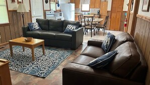 Un salon chaleureux avec un canapé, une table basse et un tapis à motifs. On aperçoit à l’arrière-plan une cuisine avec des meubles en bois et un coin repas comprenant des chaises et une table.