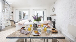 En frukostplats inredd i ett ljust och öppet vardagsrum med ett träbord, frukosttillbehör och utsikt över omgivningen.