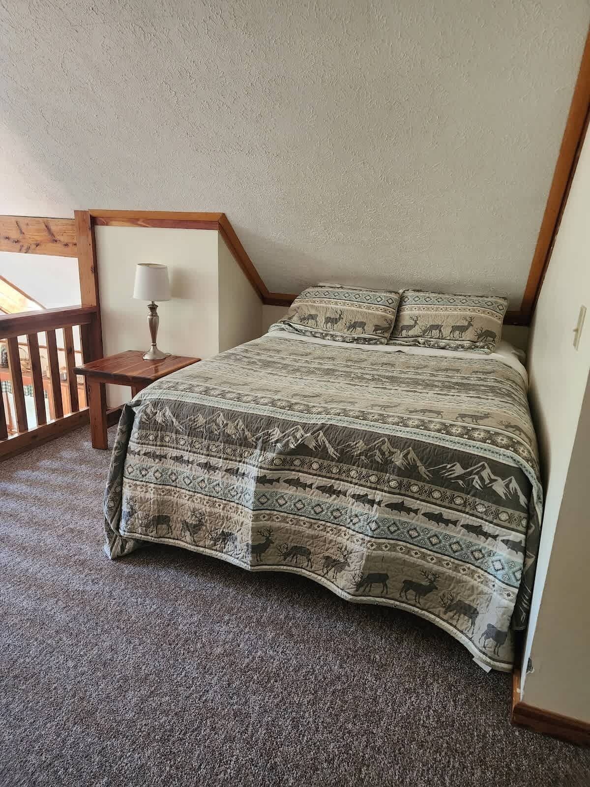 A bedroom with a bed, a nightstand, a lamp, and a view of the outdoors.