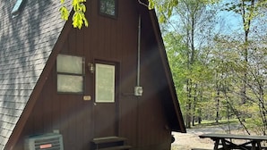 Una cabaña de madera pequeña y triangular con techo inclinado, una ventana y una puerta. Hay una mesa de picnic cerca.