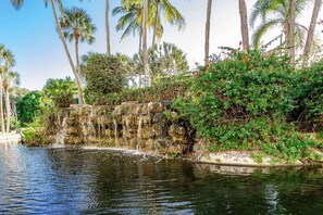 A natural water feature with cascading rocks and lush greenery, including palm trees and flowering plants. - Harbor Towers 3rd Fl 1 Bedroom Pool View (Siesta Key)