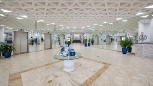 Um lobby amplo com design geométrico no teto, elevadores e plantas em vasos.