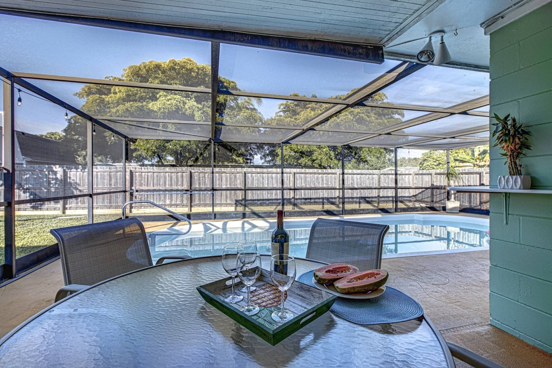 A covered outdoor area with a pool, a table with glasses and watermelon, and a view of a wooden fence and trees.