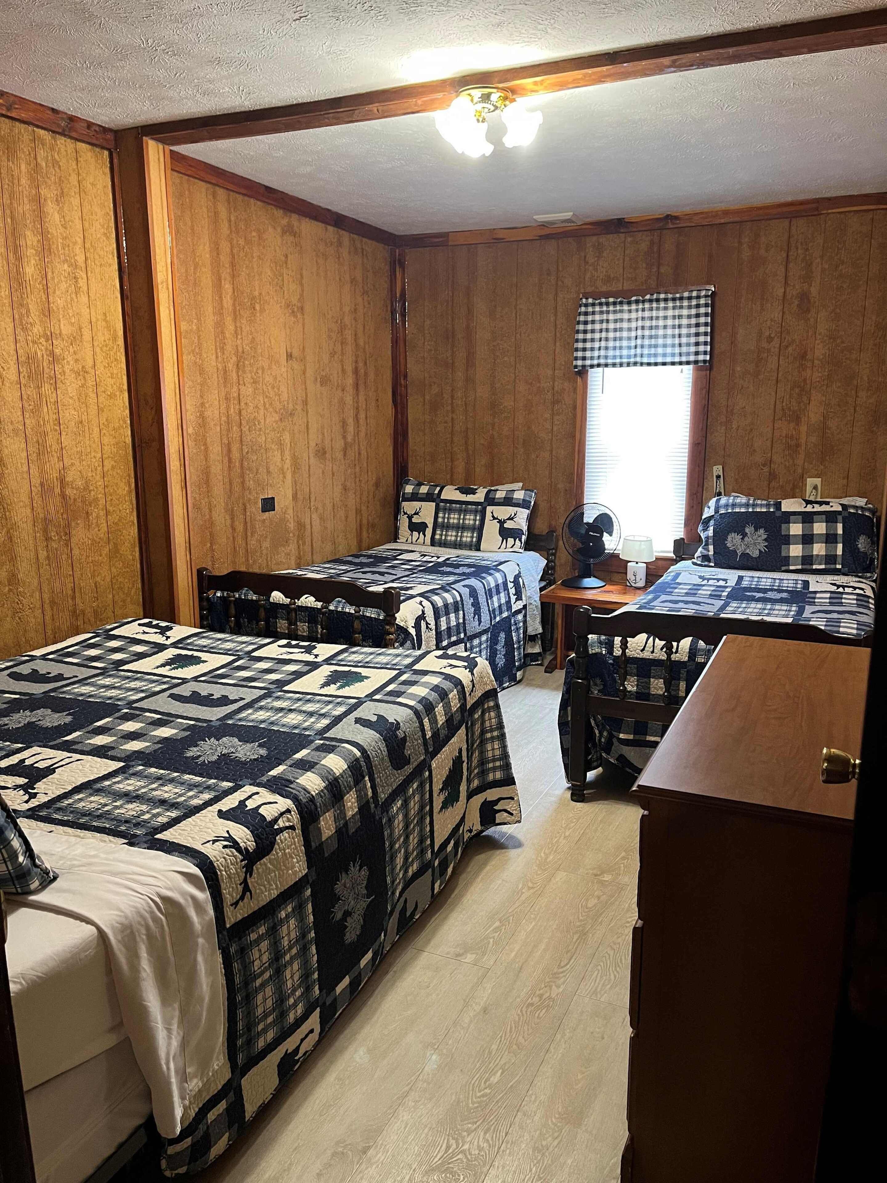 Una habitación con dos camas, paredes de madera, una ventana con persianas, un ventilador de techo y una pequeña mesa con lámpara.