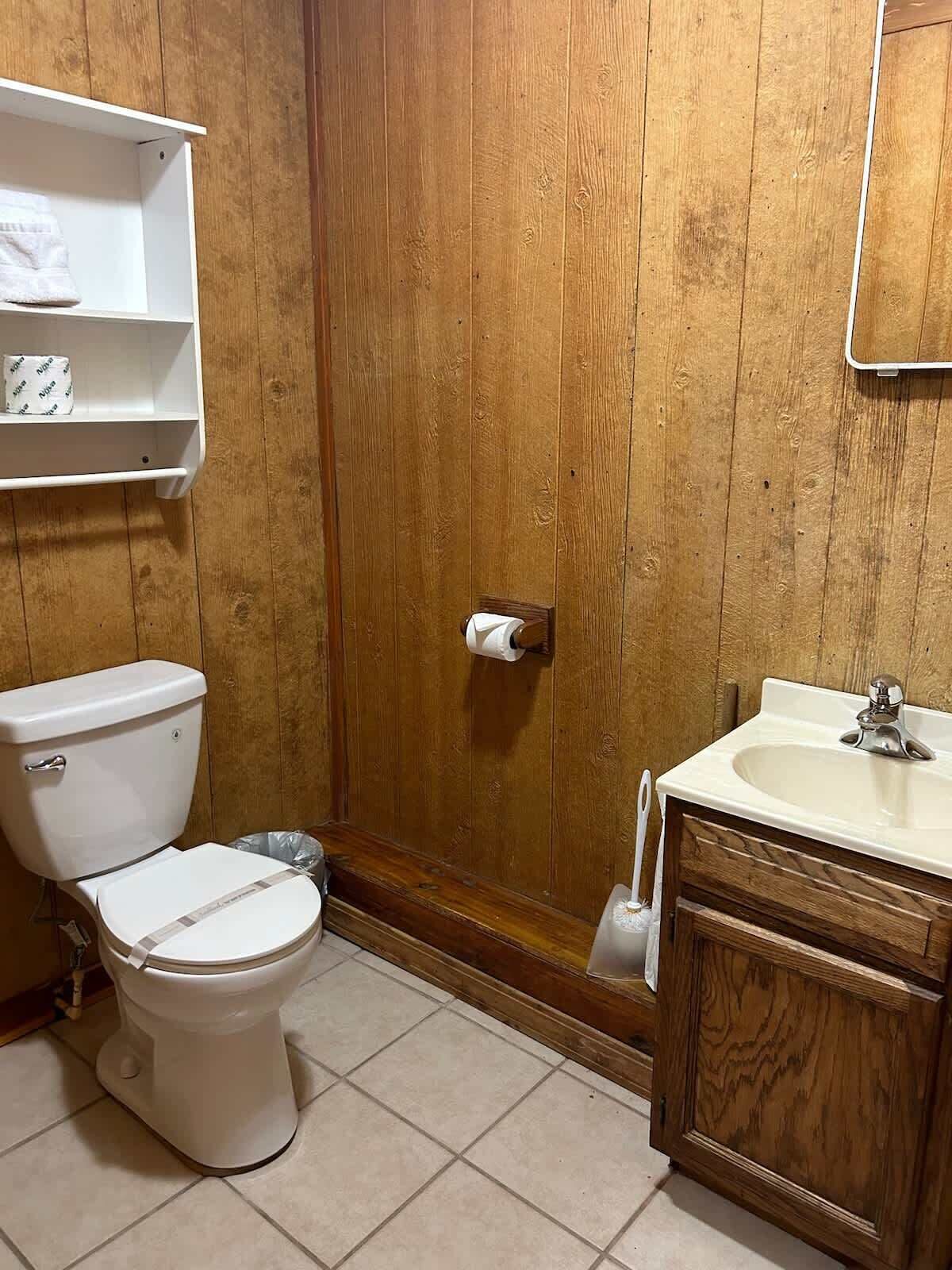 A bathroom with a wooden door, a toilet, a sink, and a shelf with toiletries.