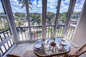A balcony with a glass table set for dining, overlooking a tropical landscape with palm trees and a pool.