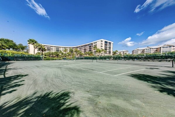 A tennis court with a net, surrounded by a fence, and buildings in the background. - Harbor Tower Gulf Luxe Escape (Siesta Key)