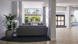 A modern lobby with a grey sofa, a large potted plant, and a view of palm trees through large windows. - Harbor Towers 4th Fl Modern 3BR With Den (Siesta Key)