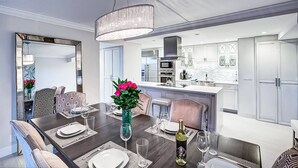 A modern dining area with a dark wooden table set for six, surrounded by patterned chairs. A large mirror reflects the room, and a chandelier hangs from the ceiling. - Harbor Towers 4th Fl Modern 3BR With Den (Siesta Key)