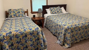 Una habitación con dos camas individuales, cada una con ropa de cama floral, una mesita de noche con lámpara, una ventana con persianas y una pequeña cesta fijada a la pared.