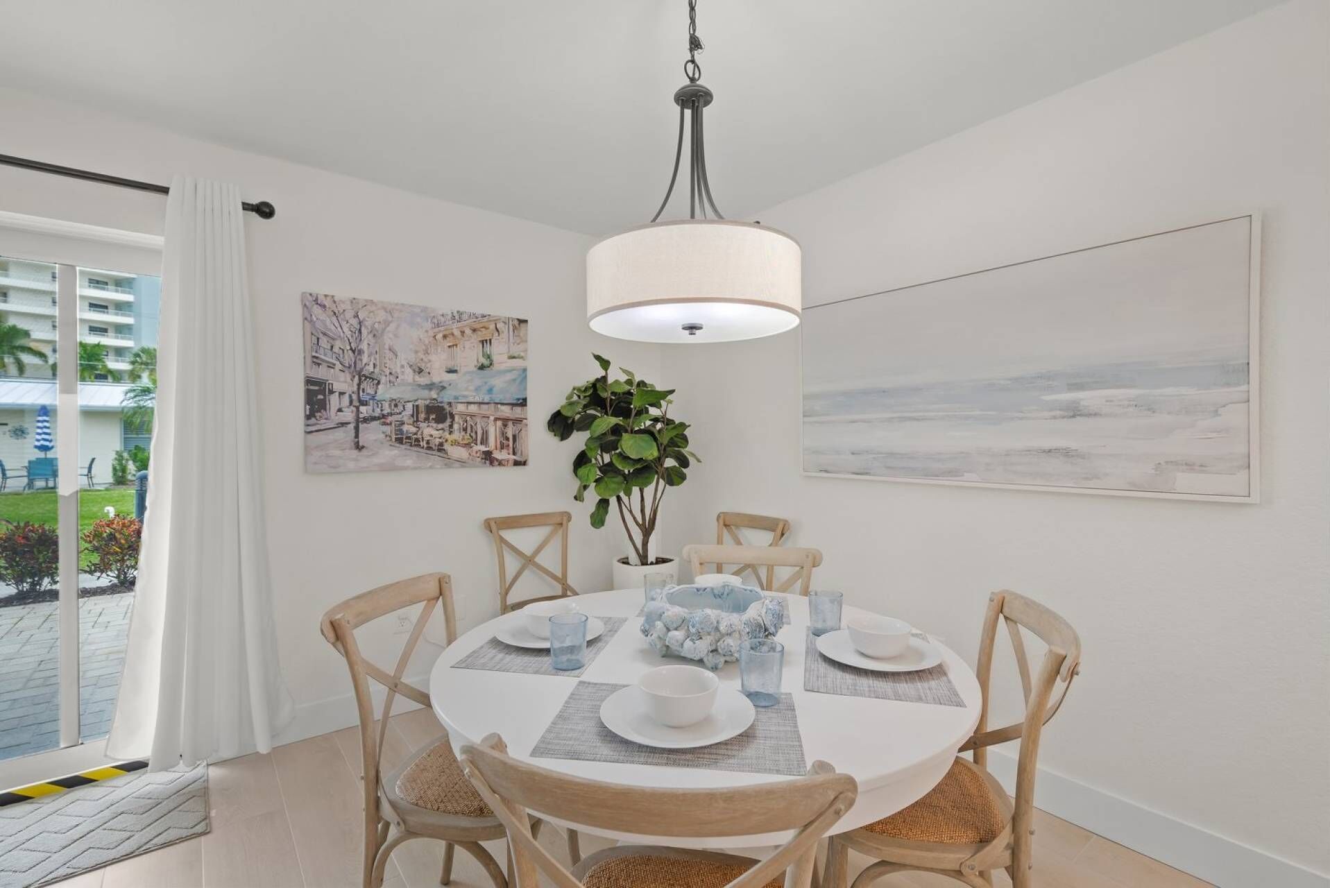 A dining area with a round table, four chairs, a hanging light fixture, and two paintings on the walls.