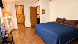 Ein Schlafzimmer mit Bett, Schreibtisch, Computer, Ventilator und einer Tür zu einem anderen Raum.
