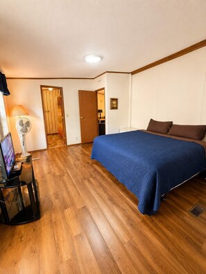 A bedroom with a bed, a desk with a computer, a fan, and a door leading to another room.
