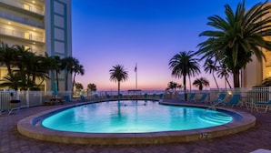 Piscina circular de 1 metro 10 de profundidad, rodeada de palmeras y sillas de descanso, con vista a un edificio al fondo.