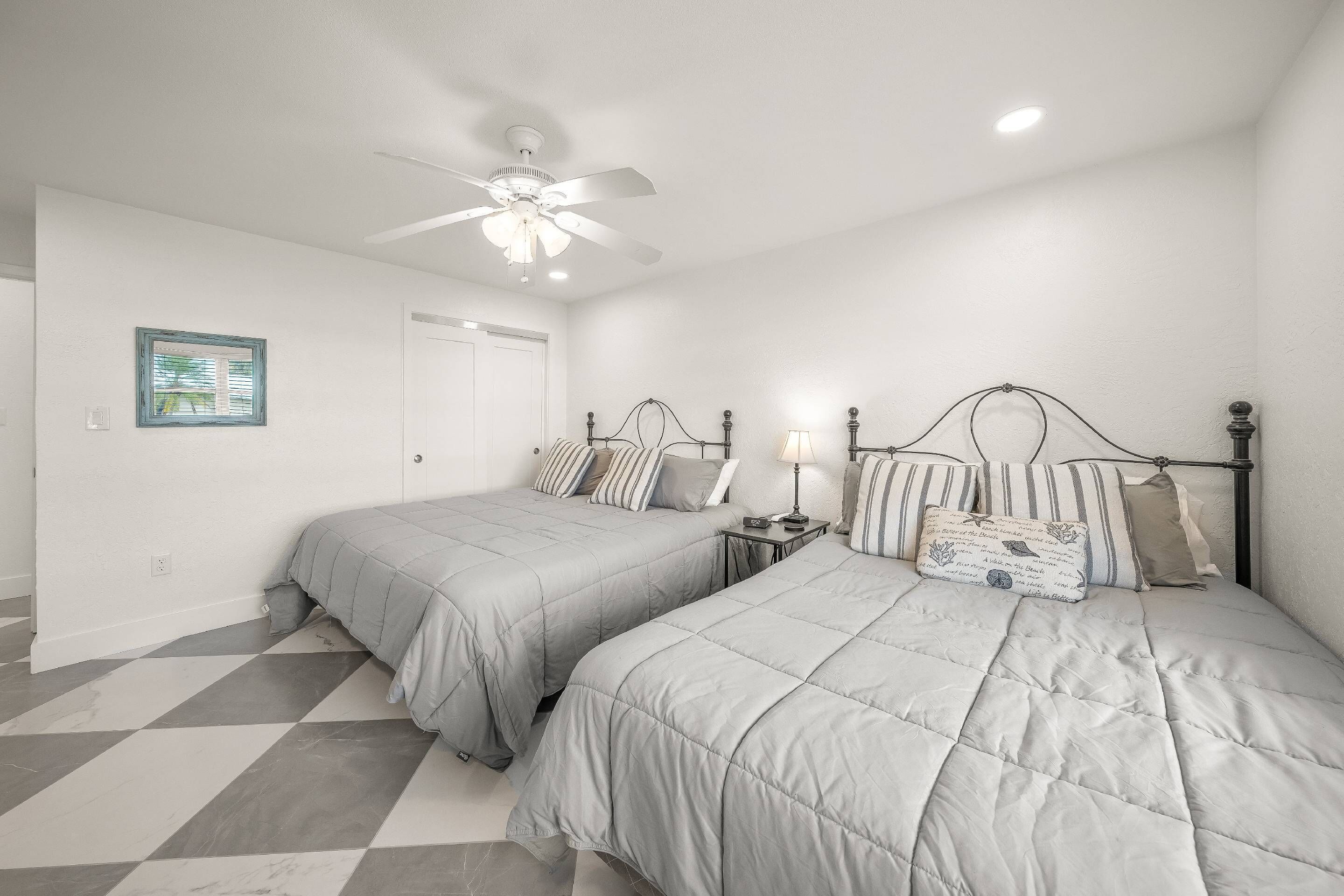 A bedroom with two beds, a ceiling fan, a nightstand with a lamp, and a window with blinds.
