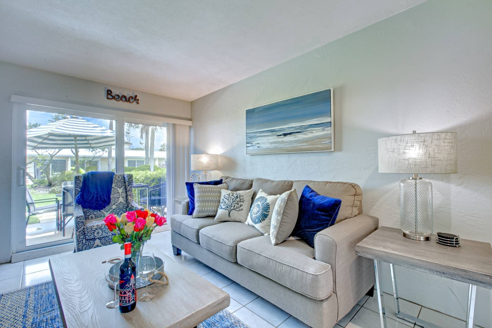 A living room with a beige sofa, a coffee table with a bottle and glasses, a painting of a beach, and a sign that reads "Beach".