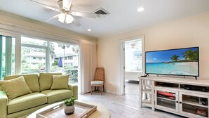 A living room with a green sofa, a coffee table with a plant, a TV on a wooden stand, and large windows with a view of a beach. - Kingston Arms 1st Floor (Sarasota)