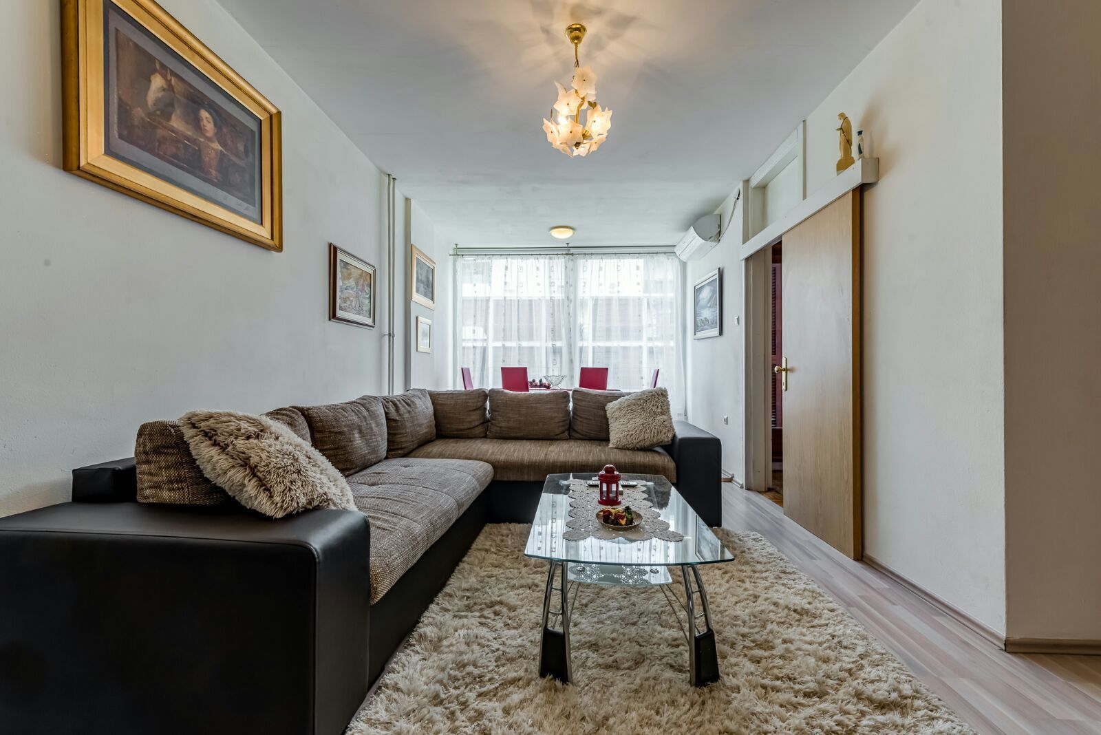 Una sala de estar con un sofá modular, una mesa de centro de vidrio y una alfombra mullida. Hay cuadros enmarcados en las paredes y un candelabro colgando del techo.