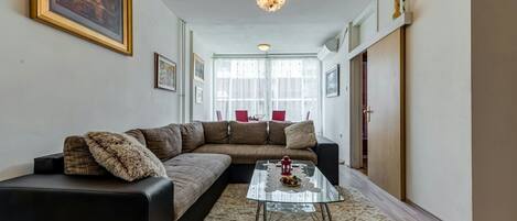 Un salon avec un canapé d’angle, une table basse en verre et un tapis moelleux. Les murs sont ornés de tableaux encadrés et un lustre est suspendu au plafond.
