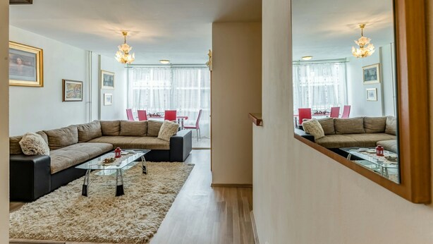 Ein modernes Wohnzimmer mit einem Ecksofa, einem Glastisch und einem flauschigen Teppich. Im Hintergrund ist eine Essecke mit roten Stühlen und einem Tisch zu sehen.