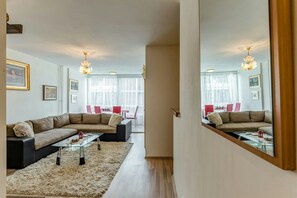 Una sala de estar moderna con un sofá modular, una mesa de centro de vidrio y una alfombra mullida. Se observa un comedor con sillas rojas y una mesa al fondo.