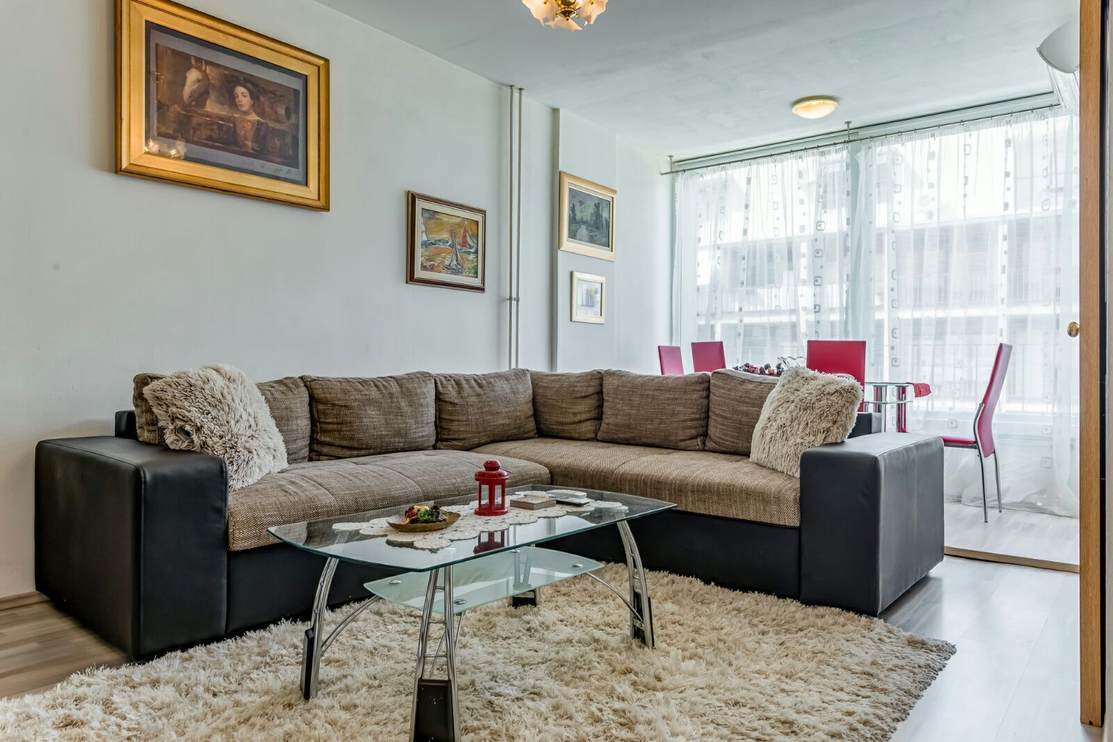 A modern living room with a sectional sofa, a glass coffee table, and a fluffy rug. There are framed artworks on the wall and a dining area with red chairs.
